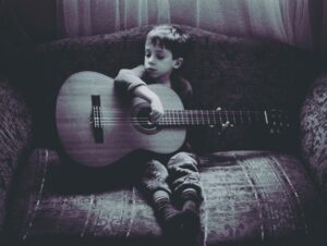 small boy sitting on sofa holding an acoustic guitar which is too big for him. His left arm is draped over the guitar and he is looking absent mindedly to his left. the photo is in black and white and looks old