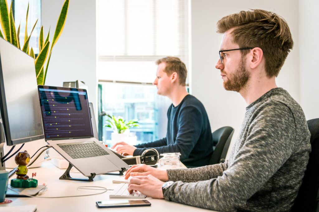 Two men sitting at laptops in an office with a bright window behind them