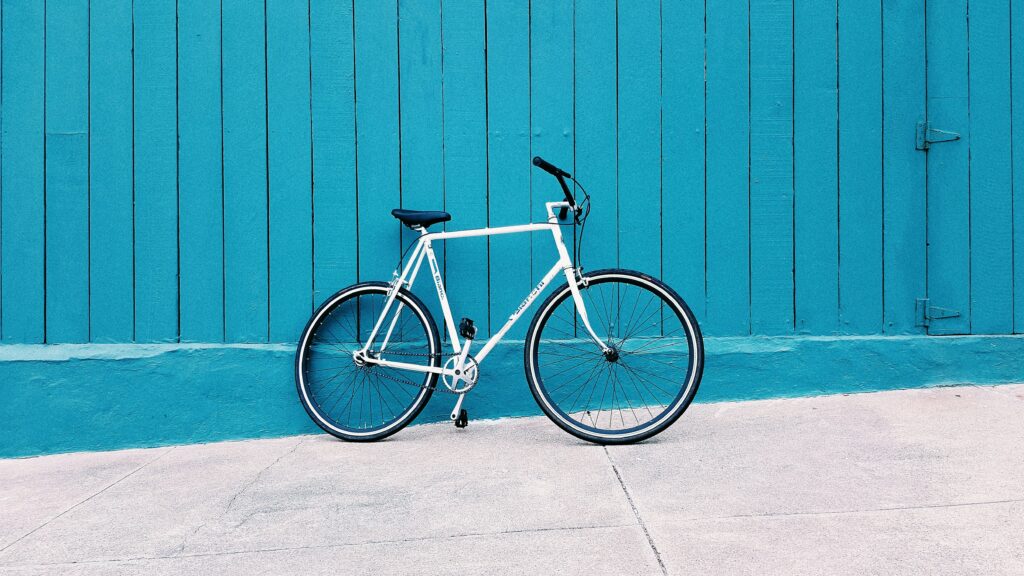 a white bicycle on a white pavement resting on a blue painted slatted wooden wall